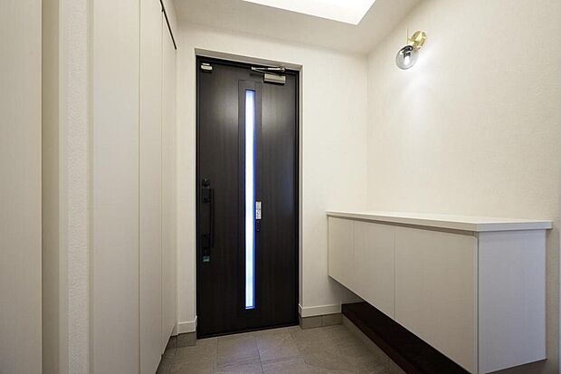 【玄関】【J-5号棟】白さ際立つ開放感あふれる玄関。スリットの窓から陽光を取り込むことで明るい空間に。壁面収納とキャビネットを設置して収納たっぷり、いつでもスッキリとした玄関回りを演出できます。