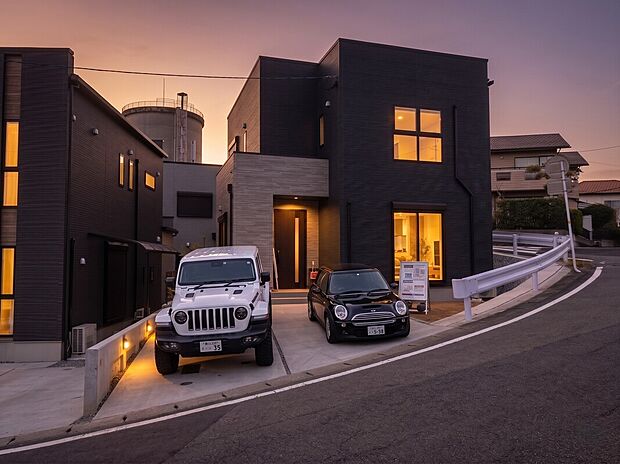 2号地外観画像：モダンな2階建ての外観が夕景に映え、ゆったりとしたコンクリートの駐車スペースに並列で2台駐車可能。大型窓から暖かな室内灯が漏れ、沿道に面した落ち着いた街並みに調和する佇まいです。
