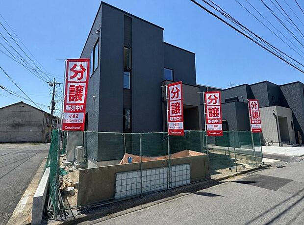 角地に建つダークトーンのモダンな2階建て外観。分譲案内の旗が並ぶ整然とした街並みで、前面は舗装道路、駐車2台分の余裕ある敷地が魅力です。周辺の落ち着いた環境が新生活の安心感を高めます。