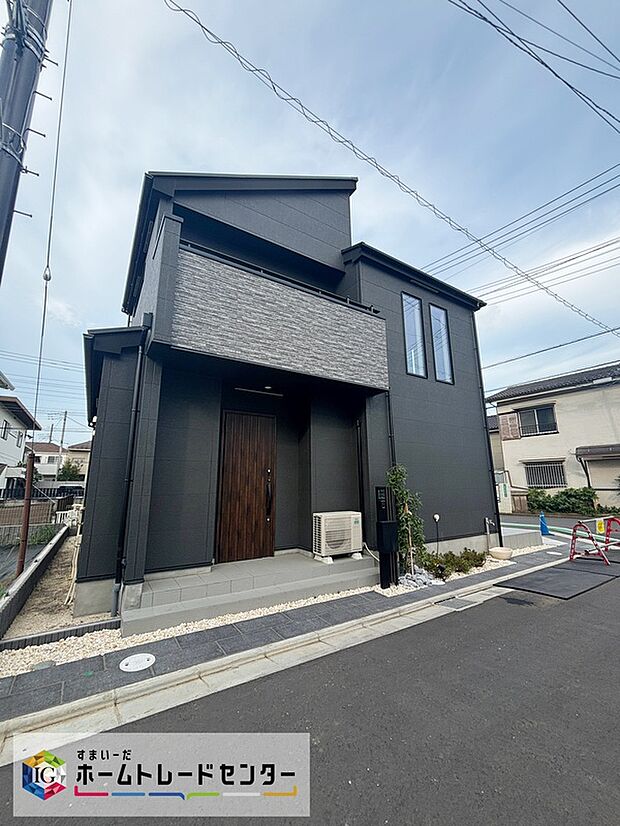 【外観(モデル棟・色・間取り変更・選ダークグレーの2階建て外観】ダークグレーの外壁と木目調の玄関扉が印象的な2階建て外観。玄関前に植栽とアプローチがあり、アスファルト舗装の分譲地内道路に面しています。