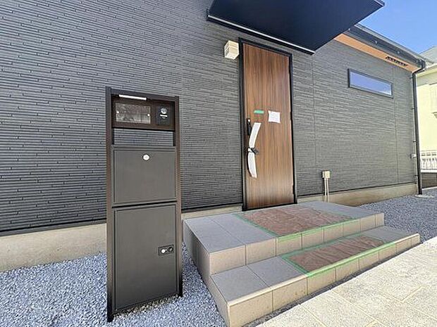 【現地外観写真】グレーの壁と木目調ドアが調和するモダンな外観。不在時でも荷物を受け取れる宅配ボックスは、子育て世帯に嬉しい設備です。