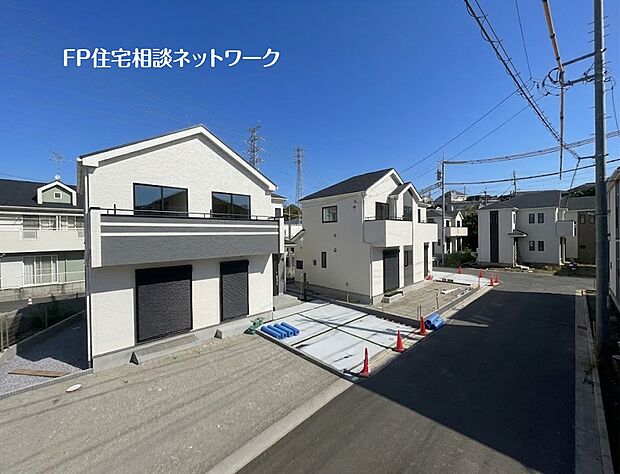 【前面道路含む現地写真】青空の下、新築一戸建て住宅が並ぶ。白い外壁に黒いバルコニーがアクセントの手前の住宅と、隣に建つ住宅。どちらもシャッター付き窓と、コンクリート舗装が施された駐車スペースがある。