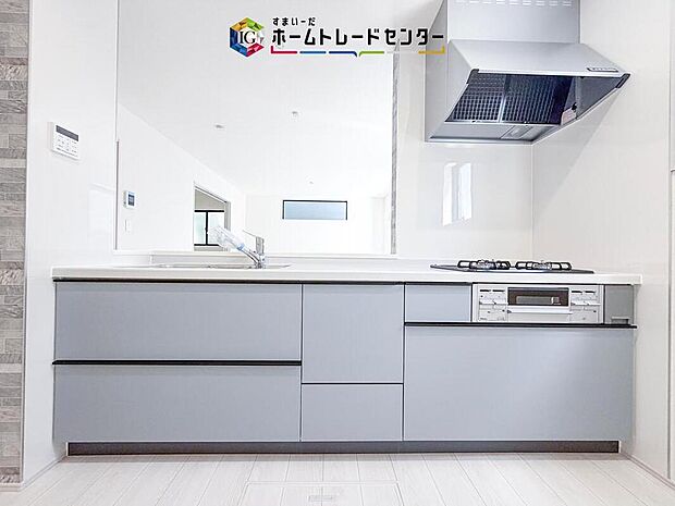 【【システムキッチン】】白いカウンタートップとシンク、ビルトイン3口コンロ、レンジフード、引き出し収納を備え、リビングを見渡せる対面配置です。