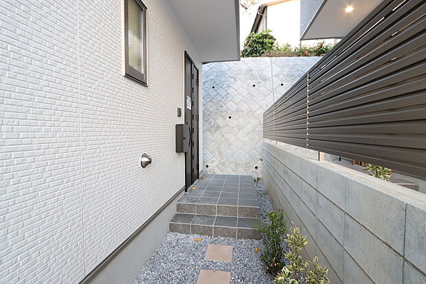 【隣接住戸との関係に配慮した玄関アプローチ　２号棟】家の顔となる玄関は、格調高いデザイン性が求められます。玄関は、高級感と断熱性、防犯性に優れた玄関ドアを標準装備。デザイン性だけではなく、ピッキング対策に優れたセキュリティサムターン等、防犯対策を考慮。