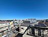 周辺：住戸からの眺望です。周辺には戸建てやマンションが建ち並び、屋根越しに街並みまで見渡せます。左手には道路と交差点が見え、奥には樹木の緑も望めます。室内とあわせて、周辺の建物との距離感もご確認ください。