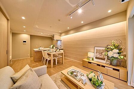 木目調のフローリングやアクセントパネルに加え、天井のダクトレール照明や壁面の間接照明、ダウンライトが配された内装で、奥には対面式のシステムキッチンを備えたリビングです。