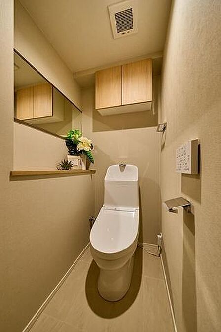 温水洗浄機能付きの便座や壁掛けリモコンを備えたトイレには、上部に木目調扉の吊戸棚がありストック品を整理できるほか、側面には横長の鏡が配置された棚付きカウンターが設置されております。