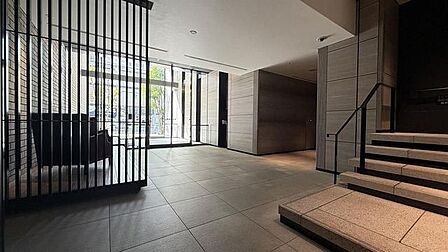 シックなトーンのタイル敷きの床と壁面で仕上げられたロビーには、奥にガラス張りの出入口が配されているほか、右手には手すり付きの階段、左側には黒色の縦格子仕切りが設置されています。