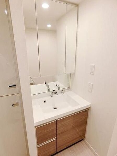 木目調の扉と清潔感のあるホワイトのカウンターを合わせた洗面化粧台には、大型の三面鏡や手元鏡が備え付けられており、下部収納に加えて左側にも作り付けの収納棚が配置されています。