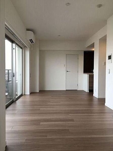 木目調のフローリングと清潔感のある白いクロスで仕上げられた洋室には、バルコニーへ出られる大きな引き違い窓やエアコンが設置されており、奥には扉や対面スタイルのキッチンが配置されています。