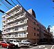 外観：白を基調としたタイル貼りの外壁と格子状の手すりが印象的な外観で、各住戸にバルコニーが設置されており、敷地内には平面駐車場や前面道路に接したアプローチが配されたマンションです。