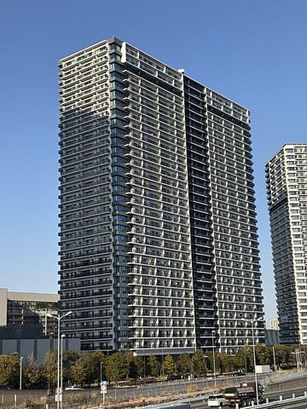 白とダークグレーを基調とした外観デザインの高層マンションで、各階のバルコニーやガラス手すりが規則正しく並んでおり、建物周辺の敷地沿いには緑豊かな植栽が整備されています。