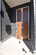 屋根付きの玄関ポーチで、雨の日の出入りもしやすく、落ち着いた外壁色と玄関ドアの木目が上品に映えます。