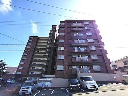 開放感のある青空の下、重厚感のあるタイル貼りの外観が目を引くマンションです。住戸数分の駐車スペースも確保されています。