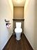 トイレ：木目調の床とアクセントクロスが落ち着いた雰囲気を醸し出すトイレ空間。清潔感のある便器に加え、便利な収納棚も備わっています。日々の暮らしを快適にする設備が整っています。