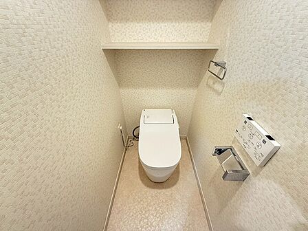 温水洗浄便座と便利な棚付きのトイレ空間。落ち着いた色合いのクロスが、リラックスできるひとときを演出します。