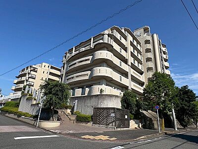 外観：青空が広がる明るい日差しの中、存在感のある佇まいが魅力のマンション。周囲の緑も豊かで、穏やかな住環境が期待できます。