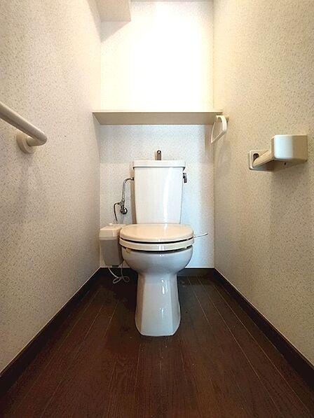 温かみのある木目調の床と清潔感のある壁紙で、落ち着いた印象のトイレ空間です。上部の棚や手すりも設置されています。