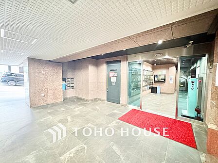 ガラス張りで明るい光が差し込む風除室には、高級感のある赤いマットが敷かれ、清潔に保たれています。 CSPのセキュリティ表示やAEDも設置されており、管理体制の良さが一目で分かります。