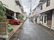 物件の南西側前面道路（北西側から）。買物施設や小中学校が徒歩10分圏内に揃う子育てにも適した住環境です。