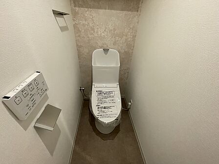 温水洗浄便座を備えた清潔感のある洗面所横のトイレ！手すりやペーパーホルダーが使いやすい位置に配置されており、機能性も良好です。壁面のシンプルなクロスが落ち着いた空間を作り出しています