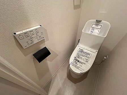 白い設備に黒いペーパーホルダーが映えるトイレ。温水洗浄便座を備え、壁付けリモコンで操作できます。手洗い付きタンクも設置され、使いやすさにも配慮。毎日使う場所を、すっきりとした印象の内装に整えています。
