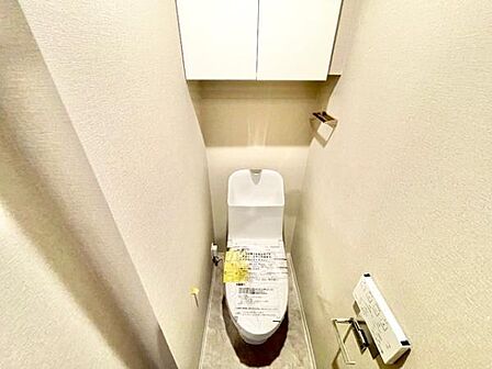 収納棚が設置されておりトイレ用品などをすっきりとまとめられます。清潔感のある内装で壁紙や床の質感も整っています。ペーパーホルダーやリモコンも近くに配置されていて便利です。