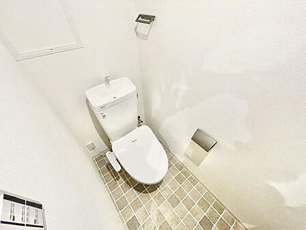 清潔感のある白を基調としたトイレです。床はタイル調でお手入れがしやすそうです。便座横にはペーパーホルダーとリモコンが設置されています。シンプルなデザインが特徴的です。