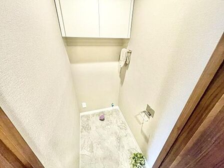 明るい色合いのクロスと床材が採用されたトイレです。上部に収納が備わっておりすっきりとした空間を実現しています。壁面にはタオル掛けやペーパーホルダーが設置されています。