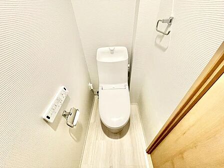 清潔感のあるトイレ空間です。壁面にはペーパーホルダーと操作パネルが設置されています。ナチュラルな木目調の床が特徴です。明るい雰囲気の内装が印象的です。コンパクトで使いやすいレイアウトです。