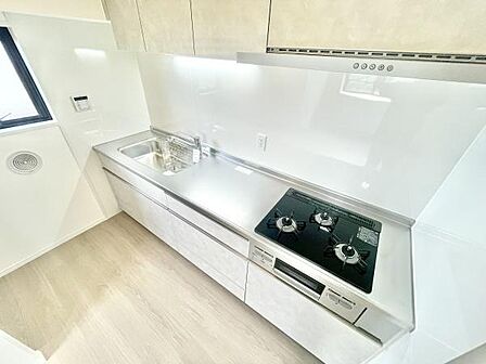 清潔感のあるキッチンは白を基調とした明るい空間です。使いやすいシンクと3口コンロが配置されています。収納スペースも豊富で調理や片付けに便利です。換気扇や窓の配置で通気性にも配慮されています。