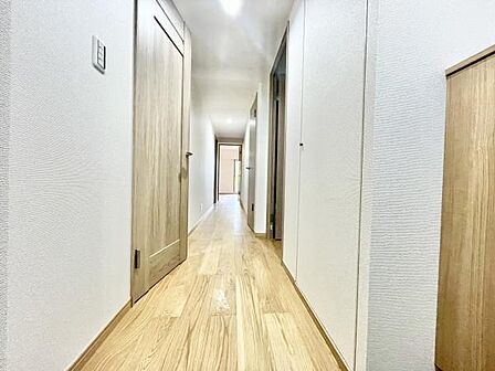 玄関から続くまっすぐな廊下が印象的です。落ち着いた木目調の床が空間を明るく演出しています。ドアや収納もシンプルで統一感があります。天井の照明が全体をしっかり照らしています。清潔感のある内装です。