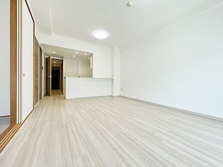明るい色合いのフローリングが特徴的なリビングです。空間が広々としておりゆとりを感じられます。ダウンライトと天井照明が室内全体を均一に照らしています。カウンター付きのキッチンスペースが見渡せる配置です。