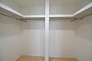 洋室7帖のWIC。大容量のL字型枕棚とハンガーパイプを完備。空間を無駄なく活用でき、衣類から季節物までスッキリ片付きます。白を基調とした清潔感あふれる明るい空間で、毎日の準備も快適に。※AI加工済み