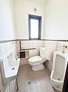 【一部AIによるモノ消し済み】窓付きで換気しやすいトイレです。個室内に手洗い場や小便器も設置されています。タイル張りで清掃しやすい設計となっています。ゆったりとした空間が確保されています。