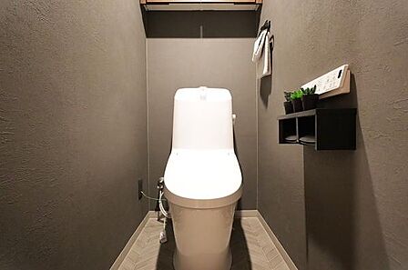 シンプルなデザインのトイレ空間です。壁の落ち着いた色合いと照明が調和しています。収納棚には植物やペーパーを置くことができます。リモコン操作で利便性を感じられます。床材のパターンも特徴的です。